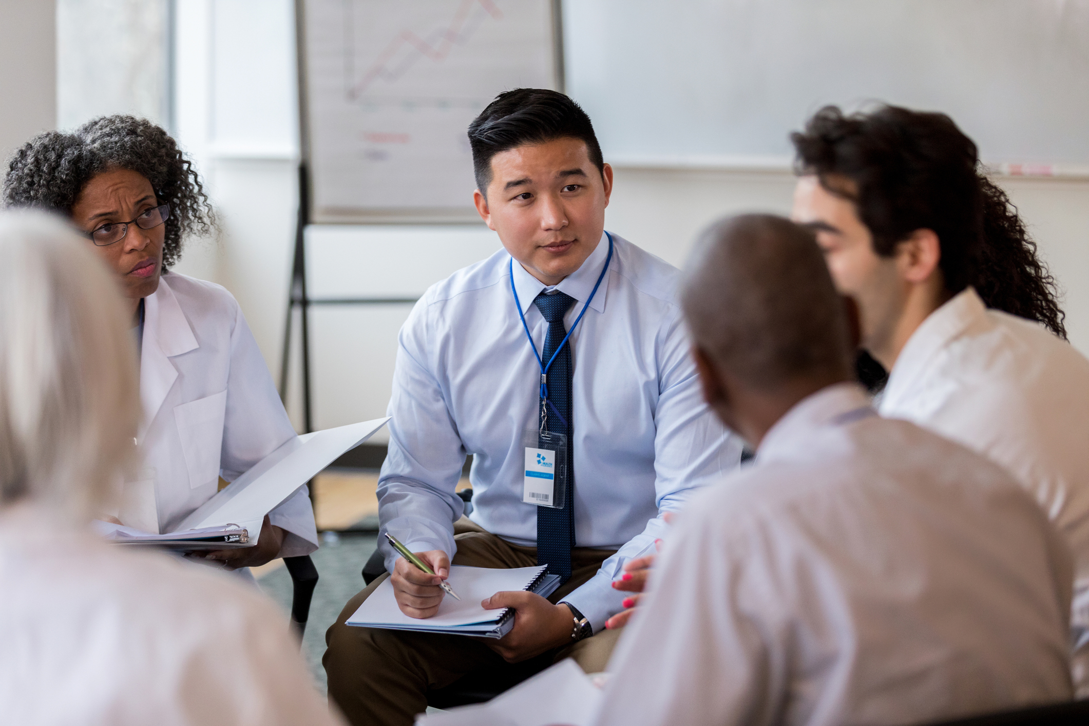 Two medical coworkers are serious as they listen to an unrecognizable coworker and take notes during a staff meeting.  They are sitting in a circle.